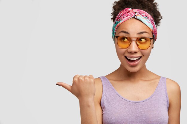 Image of glad African American female with positive expression, wears headband, points aside with thumb, has satisfied expression, sees something funny, isolated over white wall