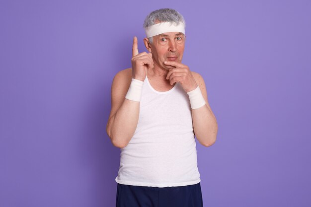 Image of good looking positive senior man standing isolated over lilac wall in , having thougthful facial expression