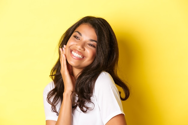 Image of gorgeous african-american woman touching her face, smiling pleased and looking left at copy space, standing over yellow background