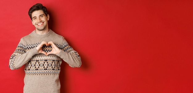Free photo image of handsome bearded guy in winter sweater wishing merry christmas and showing heart sign smiling lovely at camera standing against red background