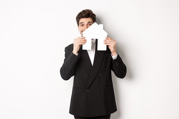 Image of handsome broker in black suit, showing house maket and smiling, selling homes, standing against white background.