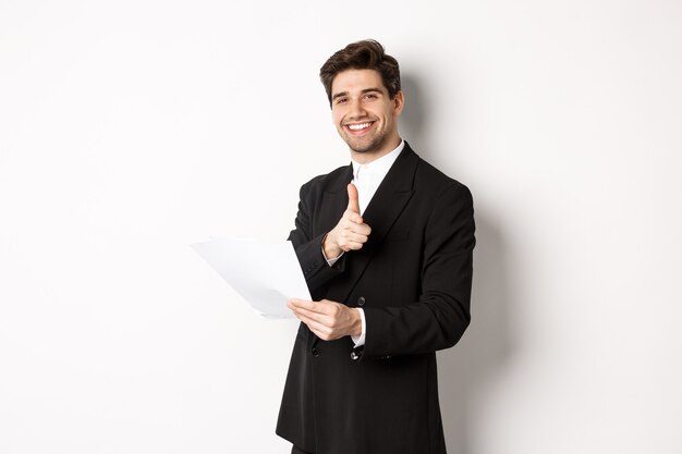 Image of handsome businessman in black suit, holding document and pointing finger at camera, praising good job, standing against white background.