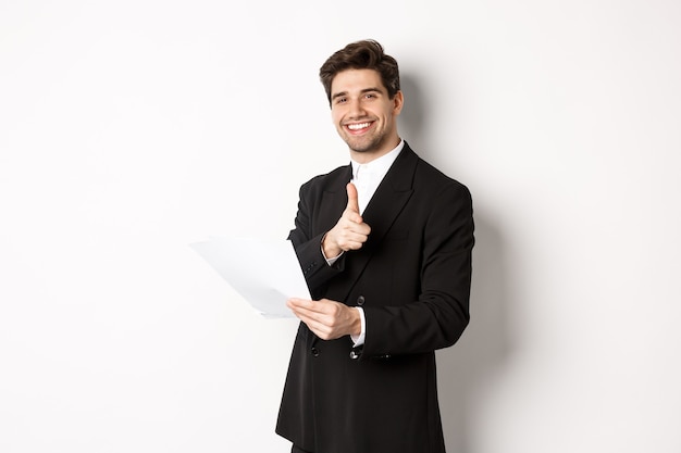 Image of handsome businessman in black suit, holding document and pointing finger at camera, praising good job, standing against white background