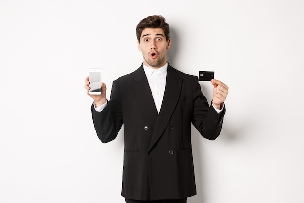 Immagine di un bell'uomo d'affari in abito nero, che sembra stupito e mostra la carta di credito con lo schermo del telefono cellulare, in piedi su sfondo bianco.