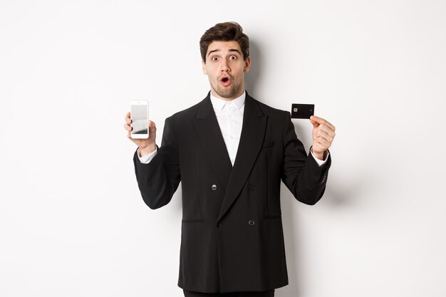 검은 양복을 입은 잘생긴 사업가의 이미지는 놀란 표정을 하고 흰색 배경에 서서 휴대폰 화면이 있는 신용 카드를 보여줍니다.