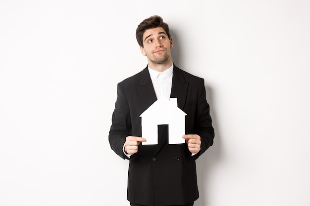 Free photo image of handsome businessman in black suit, looking for home, holding house maket and gazing dreamy at upper right corner, standing against white background