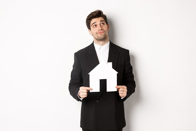Image of handsome businessman in black suit, looking for home, holding house maket and gazing dreamy at upper right corner, standing against white background
