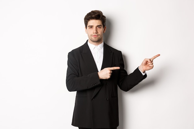 Image of handsome businessman in black suit, pointing fingers right and looking at camera, standing against white background.