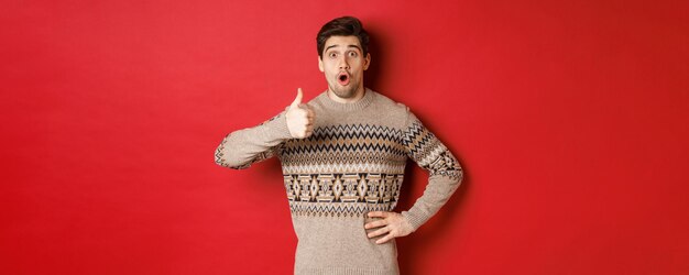 赤い背景の上に立って、何かのように、承認で親指を立てて、驚いて見えるクリスマスセーターのハンサムな白人男性の画像