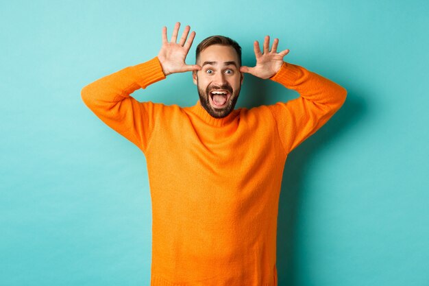 Image of handsome caucasian man making funny faces, mocking someone and smiling, standing against light turquoise wall.