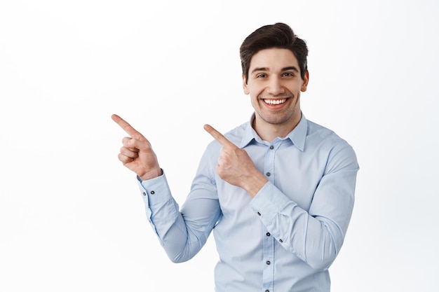 Image of handsome corporate man, businessman pointing fingers at upper left corner, showing promotional text and smiling, standing over white background