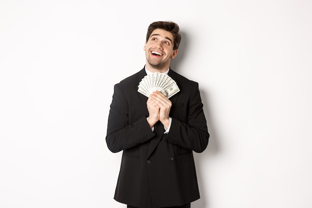 Image of handsome dreamy man in black suit, holding money and looking at upper left corner, thinking about shopping, standing over white background.