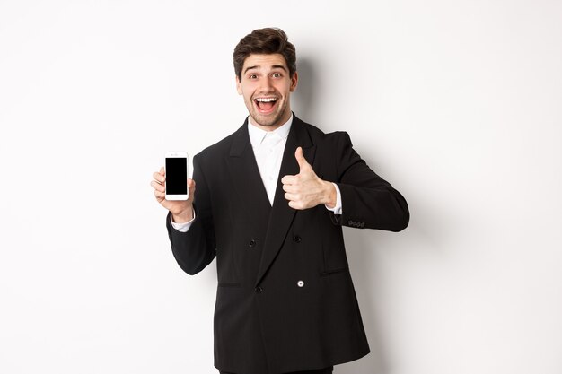 Image of handsome male entrepreneur in black suit, recommending app or online shop, showing thumbs-up and smartphone screen, standing over white background