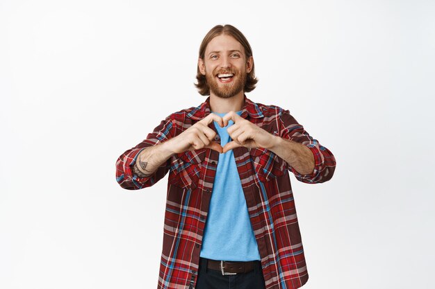 Image of handsome man, blond bearded guy showing love, heart sign and laughing, smiling cute, standing against white background.