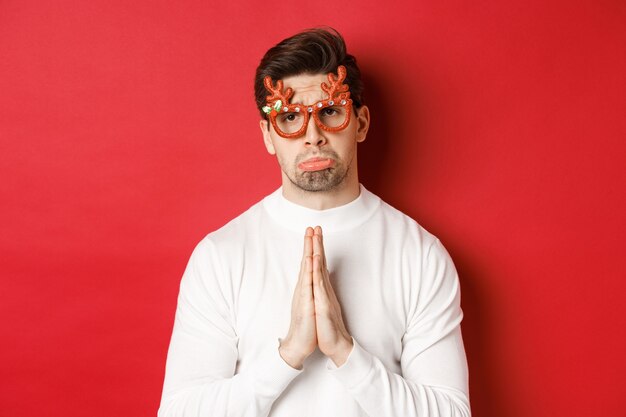 Image of handsome man in christmas party glasses, begging for help or apologizing, need a favour, standing over red background.