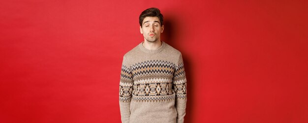 Image of handsome man in christmas sweater pucker lips and close eyes, waiting for kiss under mistletoe, standing over red background