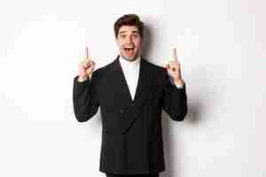 Free photo image of handsome man in party suit, showing holidays promo, pointing fingers up and smiling amazed, standing over white background