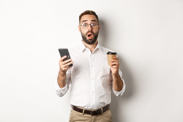 Image of handsome manage drinking coffee, reacting surprised to message on mobile phone, standing  