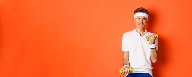 Free photo image of handsome middleaged male athlete workout with dumbbells and smiling training against orange