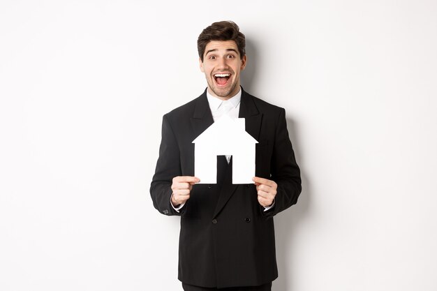 Image of handsome real estate agent in black suit showing home maket, and looking amazed, selling houses, standing against white background