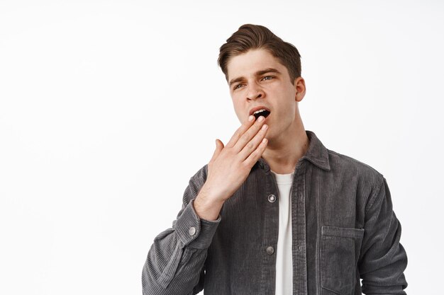 Image of handsome stylish guy, student yawning and cover opened mouth, yawn from boredom, feeling uninterested, careless and unamused, white background