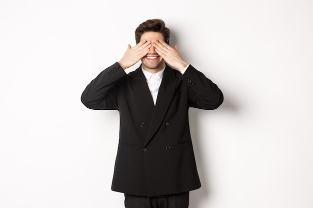 Image of handsome stylish man in black suit, waiting for christmas surprise, covering eyes with hands and smiling, anticipating presents, standing over white background