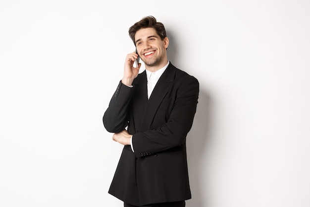 ハンサムで成功したビジネスマンが電話で話し、満足して笑って、白い背景にスーツを着て立っている画像