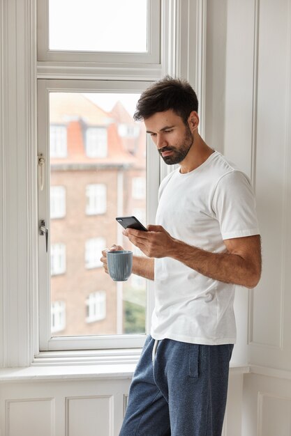 Image of handsome young Caucasian man with thick bristle, dressed in casual t shirt, uses new cellular, holds cup with coffee, stands near window