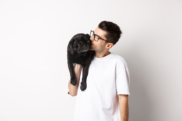 Image of handsome young man kissing his cute black pug, holding dog on shoulder, standing over white background