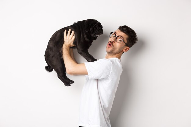 Image of handsome young man loving his pug. Dog owner holding puppy and smiling happy at camera, standing over white background.