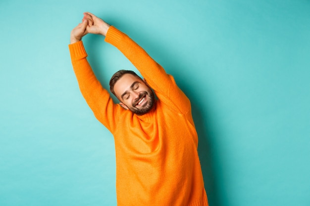 Image of handsome young man stretching hands and smiling after good rest, standing in orange sweater over light turquoise wall.