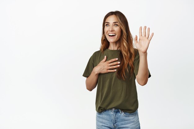 Image of happy adult woman raising one hand, place arm on heart, making promise, introduce herself, telling her name, making sincere confession, white background.