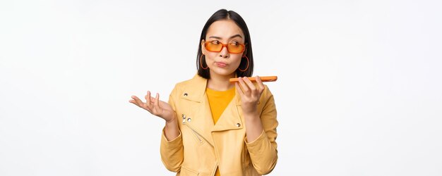 Image of happy asian girl talking on speakerphone recording translating her voice with mobile phone app talking in smartphone dynamic standing over white background