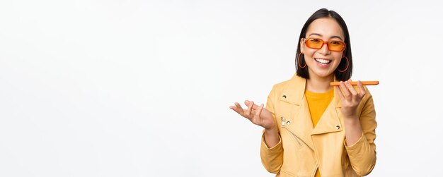 Image of happy asian girl talking on speakerphone recording translating her voice with mobile phone app talking in smartphone dynamic standing over white background