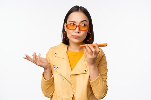 Image of happy asian girl talking on speakerphone recording translating her voice with mobile phone app talking in smartphone dynamic standing over white background