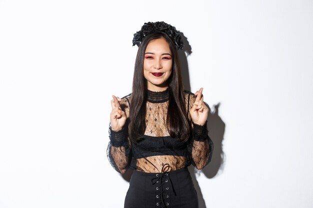 魔女に扮したハロウィーンを祝うのが大好きな幸せなアジアの女の子の画像、幸運のために希望に満ちた笑顔とクロスフィンガー、願い事をし、白い背景の上に立っています。