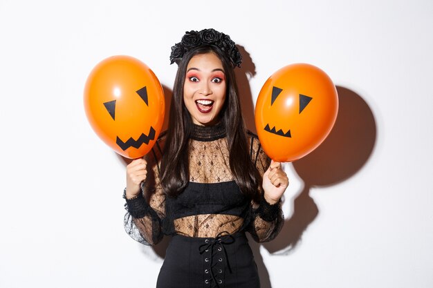 Изображение счастливой азиатской женщины в костюме ведьмы, празднующей хеллоуин, держа воздушные шары с страшными лицами, стоя над белой предпосылкой.