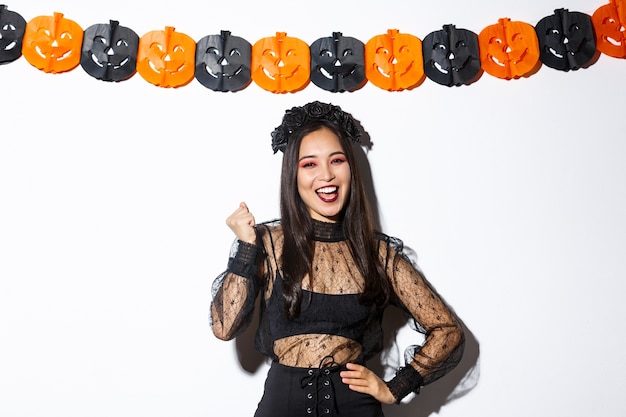 Изображение счастливой азиатской женщины в костюме ведьмы, весело проводящей время на вечеринке в честь Хэллоуина, говоря «да», жизнерадостной, стоя против баннеров тыквы.