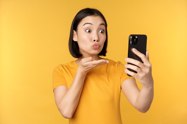 黄色の背景に立っているスマートフォンアプリケーションで話している幸せな美しいアジアの女の子のビデオチャットの画像コピースペース