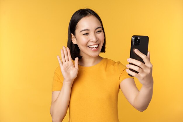 Image of happy beautiful asian girl video chatting talking on smartphone application standing against yellow background