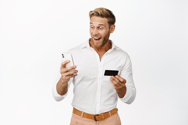 Image of happy blond man looking at his phone holding credit card order something staring surprised at application interface white background