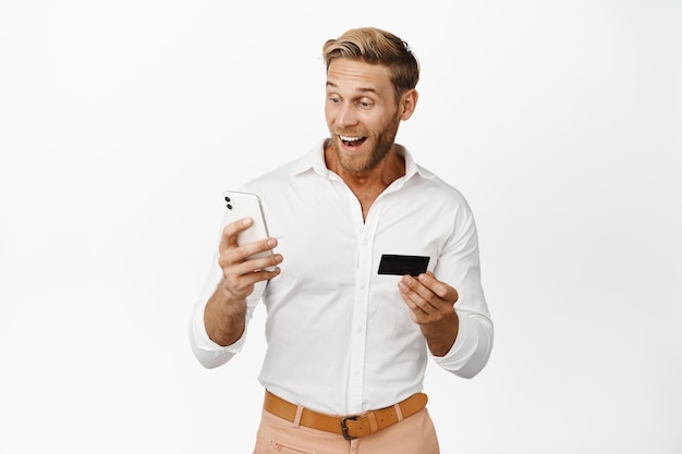 Image of happy blond man looking at his phone holding credit card order something staring surprised at application interface white background