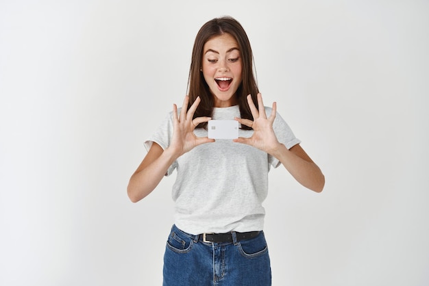 Image of happy brunette woman showing plastic credit card and looking at it with joy and amazement standing in tshirt against white background