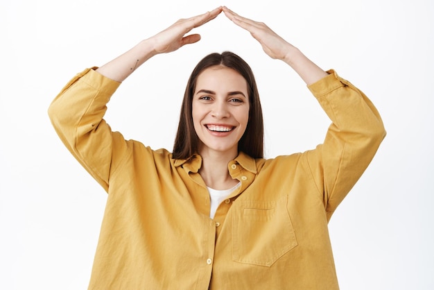 Immagine di una donna candida felice che ride sorridente e mostra un tetto sicuro che fa il tetto con le mani sopra la testa protette in piedi su sfondo bianco