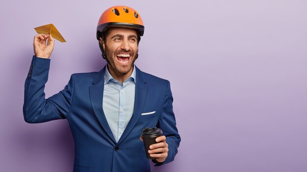 Image of happy carefree male engineer throws handmade plane, drinks coffee, glad to achieve success at work, looks into distance and laughs