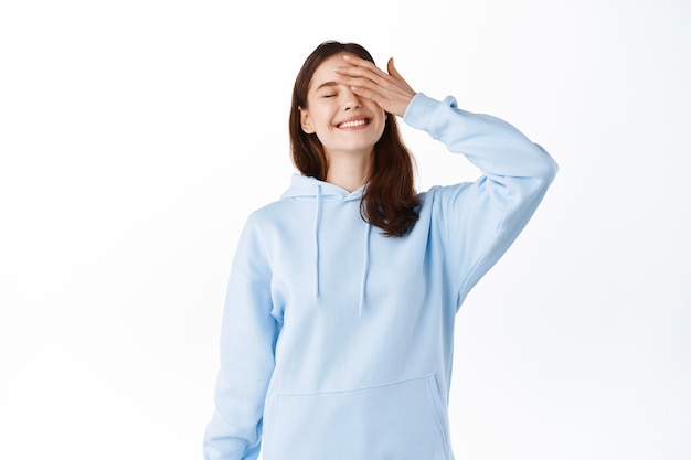 Image of happy and carefree teen girl smiling and touching face with hand, close eyes joyfully, feeling relaxed and upbeat, standing against white wall