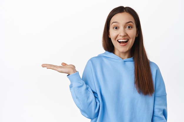Image of happy cute girl holding in open hand, smiling excited about product on sale, showing item on her palm, empty space, standing against white background