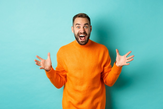 Image of happy and excited man announce big news, spread hands and shouting of joy, rejoicing, standing over light turquoise wall.
