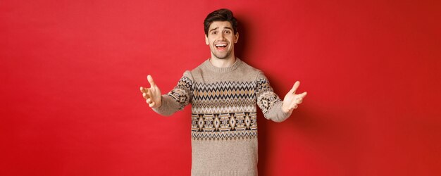 Image of happy and flattered handsome guy in christmas sweater, reaching hands to welcome guests on new year party, inviting to celebration, standing over red background