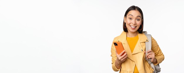 Image of happy girl traveller tourist with backpack looking at smartphone using mobile phone route app standing over white background
